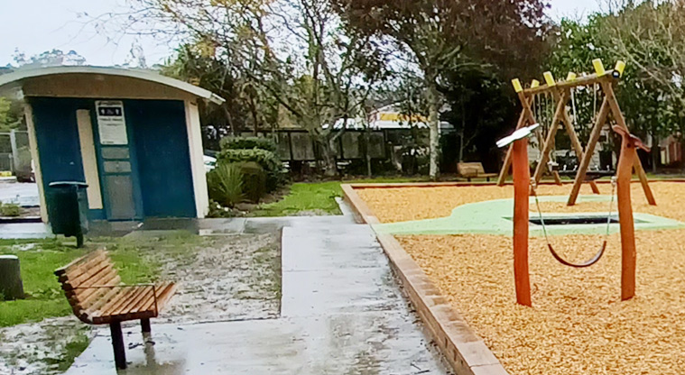Collins Park - Accessible toilet block with a footpath and the swings on the right, and seating in the foreground.