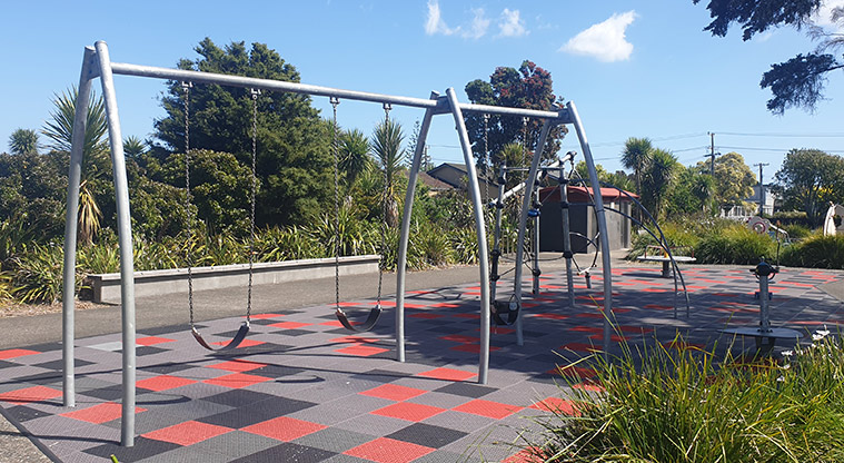 Kuaka Park - Set of three swings with climbing and spinning equipment, toilets and trees in the background. Photo credit: J Grigg.