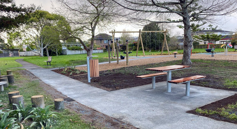 Maygrove Park – Section of footpath along the edge of the playground with a picnic table and seating, rubbish bin, trees, and the playground in the background.