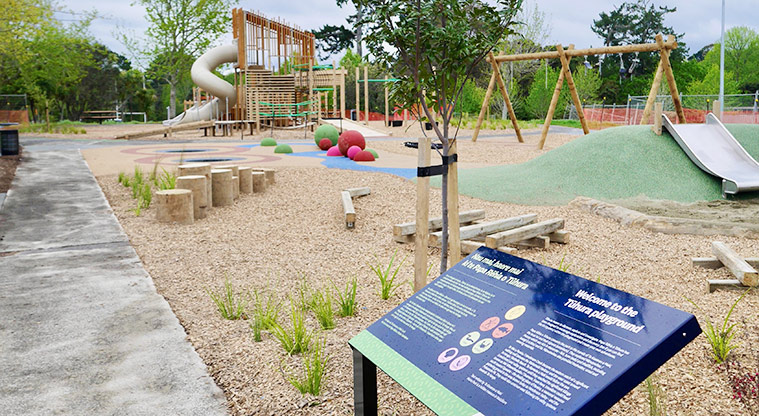 Moire Park - Section of path with a sign describing the playground, balance logs and poles on a bark base, and the rest of the playground in the background.