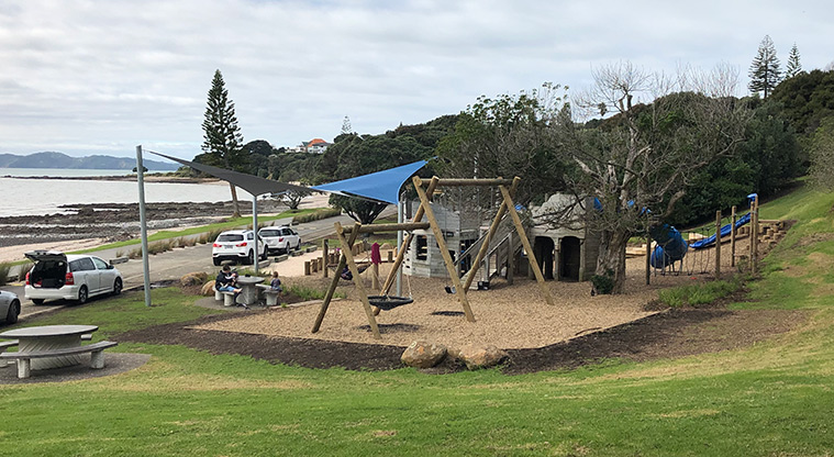 Omana Esplanade - The playground with a picnic table, the road and view of the Hauraki Gulf Islands.