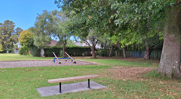 Orchard Reserve - A bench seat beside a large tree with the seesaws in the background. Photo credit: T Hodder.