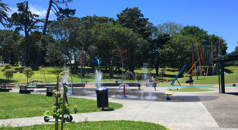 Potters Park – Splash pad and playground with a small hill and trees in the background.