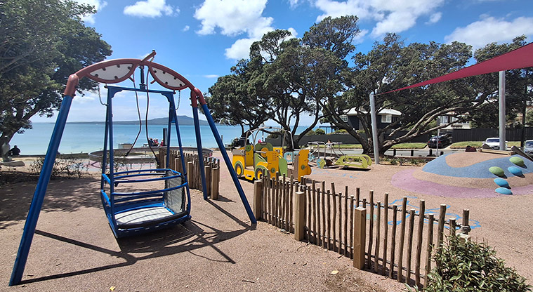 Takapuna Beach Reserve - Section of the playground with a self-propelled swing, rocking truck and a play mound and swings under a red shade sail.