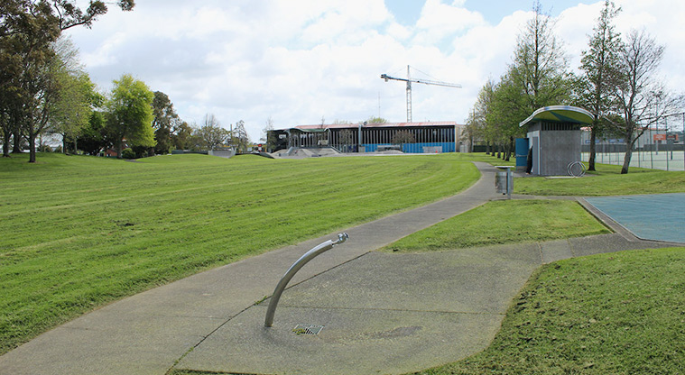 Te Pai Park - Path with the drinking fountain and toilets on the right, and open space and trees on the left.