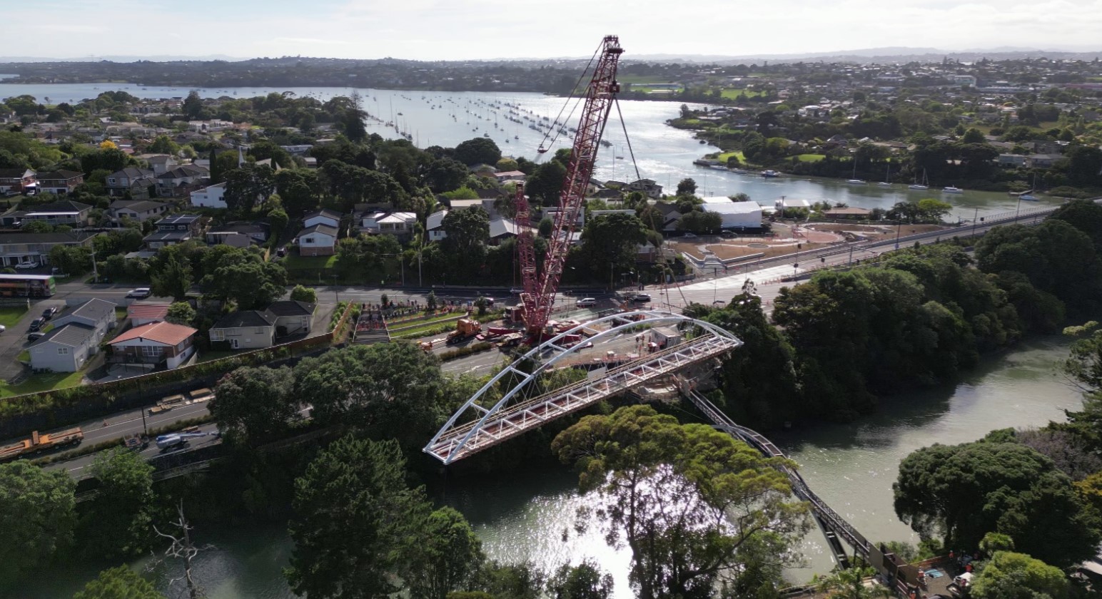 The new three-metre-wide white steel arch bridge crossing Panmure Basin being lifted into place by a red crane with the estuary, houses and greenery in the background.