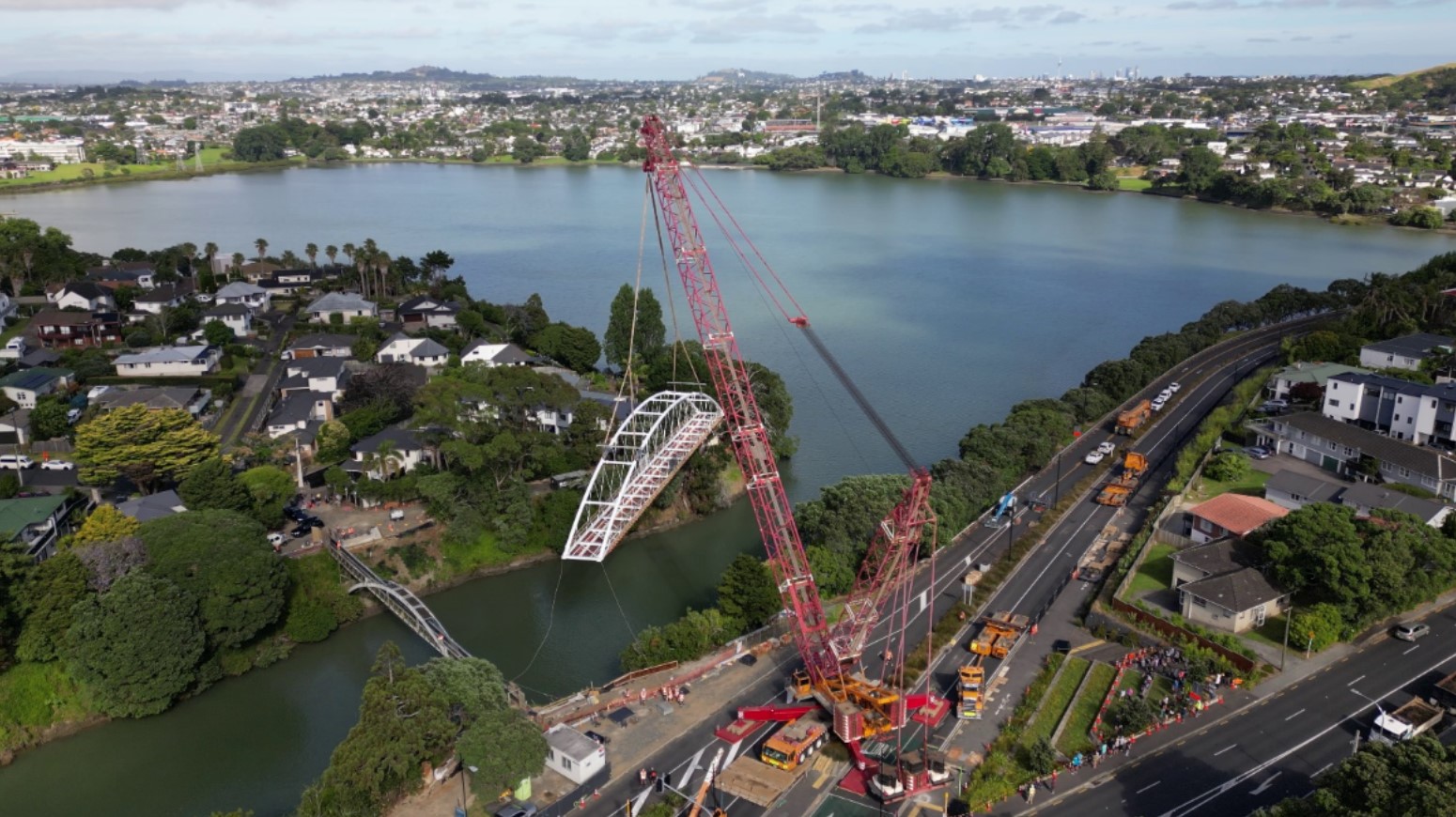 The new three-metre-wide white steel arch bridge crossing Panmure Basin being lifted into place by a red crane with the estuary, houses and greenery in the background.