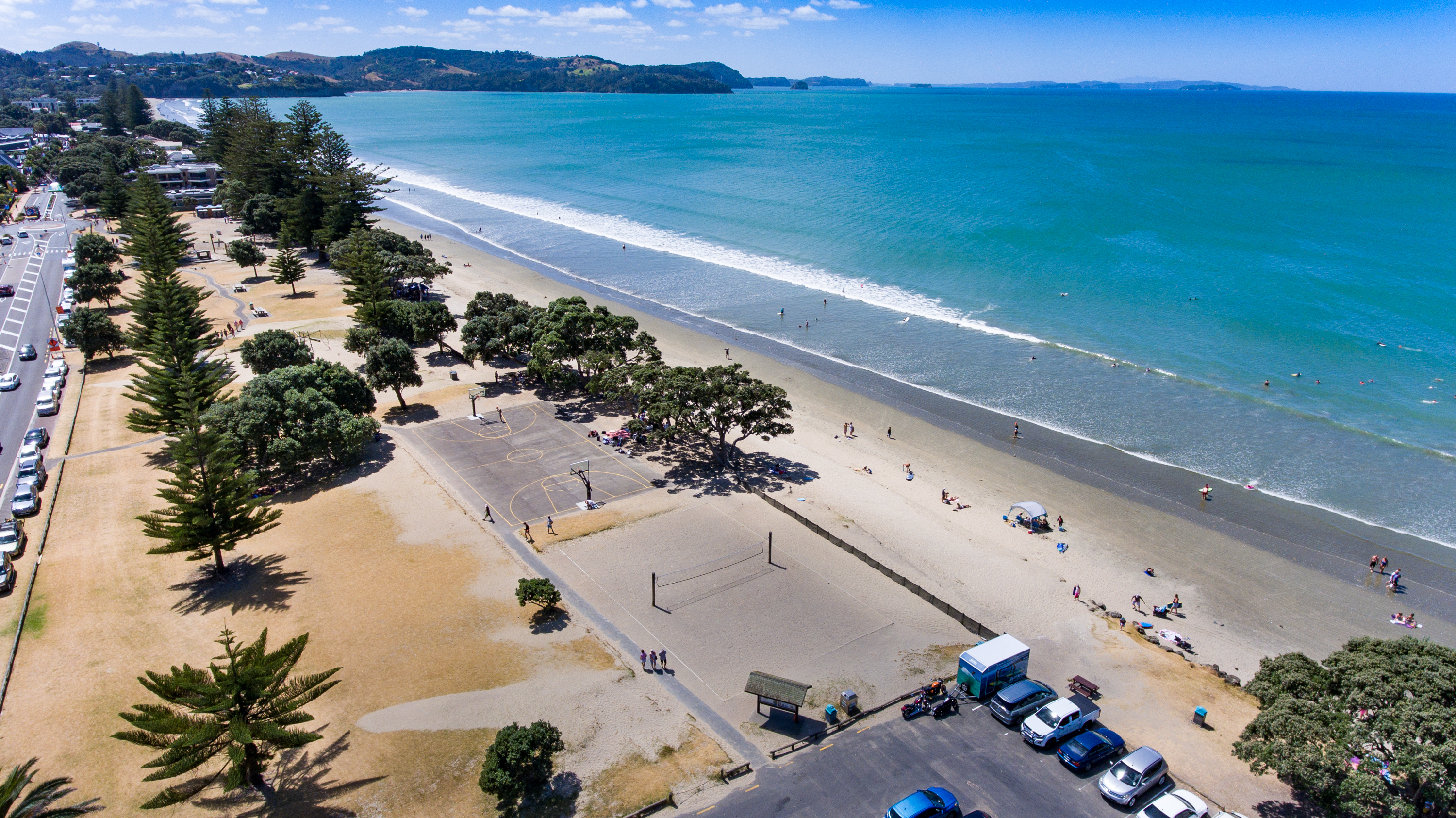 Section of Ōrewa Reserve with trees, basketball and volleyball court and car parking. People are walking, standing, lying and swimming on the beach.